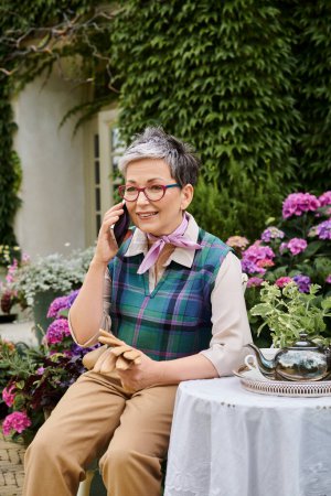 mature cheerful woman sitting in garden at tea time and talking by phone near house in England