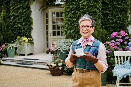 elegant jolly mature woman in chic attire drinking tea near house in England and smiling at camera