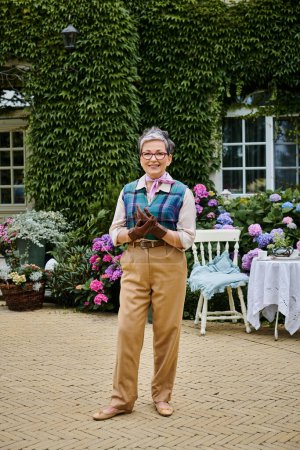 élégante femme gaie mature en tenue chic posant près de sa maison en Angleterre et regardant la caméra