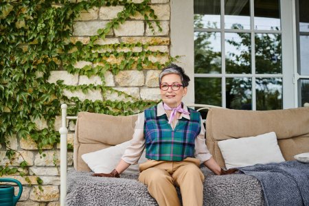 attraktive fröhliche reife Frau sitzt auf Sofa in der Nähe ihres Hauses in England und schaut in die Kamera
