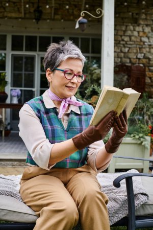 kultivierte reife lebenslustige Frau mit Brille liest Buch in der Nähe ihres Hauses in ländlichem England