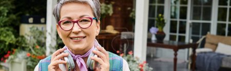 Téléchargez les photos : Debonair femme mûre dans des lunettes élégantes posant avec des écouteurs et souriant à la caméra, bannière - en image libre de droit