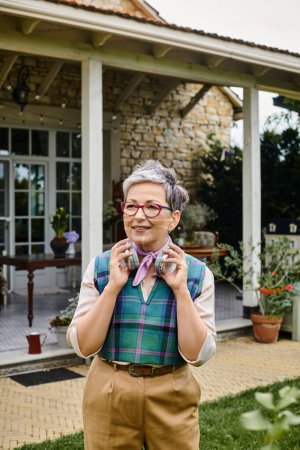 mujer madura alegre debonair en traje elegante con auriculares mirando hacia otro lado cerca de su casa en Inglaterra