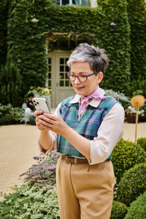 kultivierte fröhliche reife Frau in schicker Kleidung, die in der Nähe ihres Hauses in England auf ihr Smartphone schaut