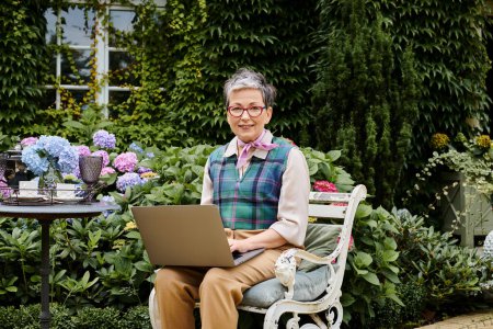 elegant cheerful mature woman with short hair sitting with laptop at tea time and smiling at camera