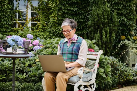 Téléchargez les photos : Debonair mature joyeuse femme en tenue chic assis avec ordinateur portable à l'heure du thé près de sa maison en Angleterre - en image libre de droit
