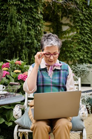 raffiné mature joyeuse femme en tenue chic assis avec ordinateur portable à l'heure du thé près de sa maison en Angleterre