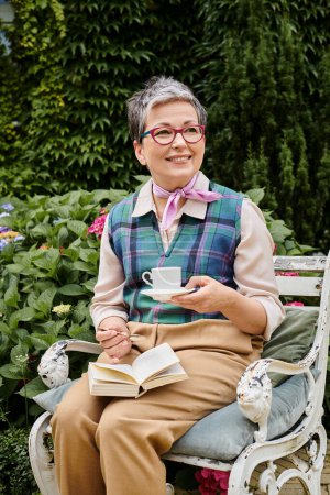 Téléchargez les photos : Raffiné mature femme gaie avec des lunettes profiter de thé chaud et réserver près de sa maison en Angleterre - en image libre de droit