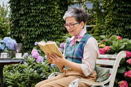 elegante, reife, fröhliche Frau mit Brille und Lesebuch zur Teezeit in der Nähe ihres Hauses im ländlichen England