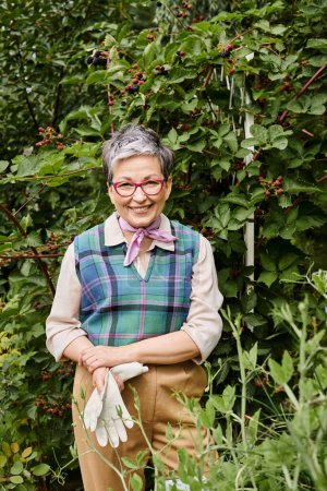 joyful refined mature woman with gloves and glasses taking care of her plants and smiling at camera