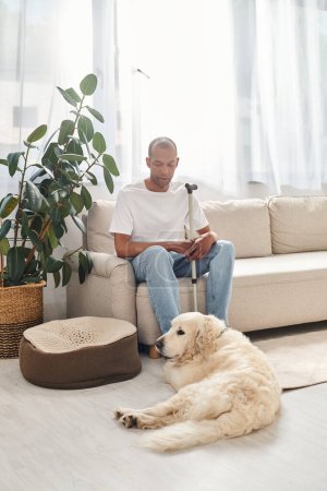 Téléchargez les photos : Homme afro-américain handicapé avec myasthénie gravis assis à côté de son chien Labrador fidèle sur un canapé confortable à la maison. - en image libre de droit