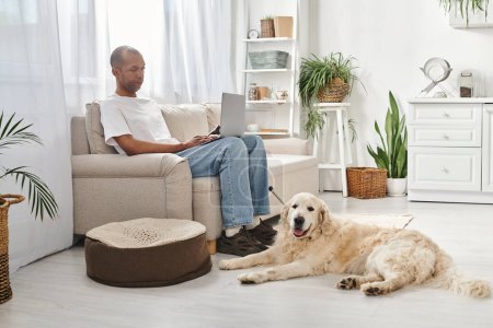 Foto de Un hombre afroamericano con miastenia gravis se sienta en un sofá, usando una computadora portátil con su perro Labrador a su lado en casa. - Imagen libre de derechos