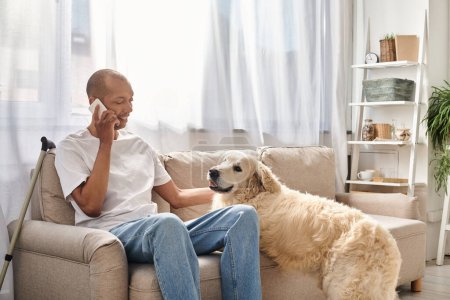 Un hombre afroamericano, discapacitado con miastenia gravis, se sienta en un sofá hablando en un teléfono celular junto a su leal perro Labrador.