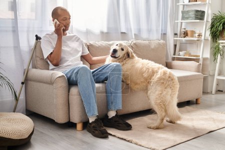 Un hombre afroamericano se sienta en un sofá, hablando en un teléfono celular junto a su leal perro Labrador en casa.