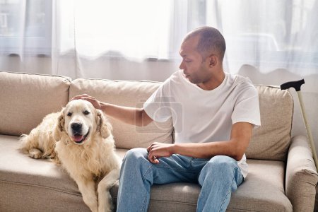 A man, disabled with myasthenia gravis, sits on a sofa petting a Labrador dog, showcasing diversity and inclusion.