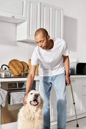 Foto de Un hombre afroamericano con miastenia gravis acaricia pacíficamente a su perro labrador en una acogedora cocina. - Imagen libre de derechos