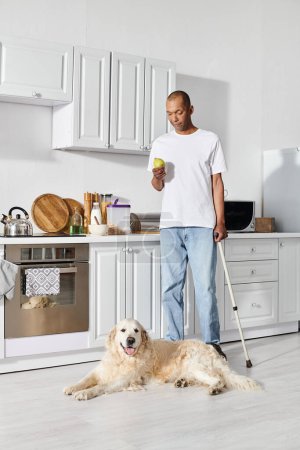 Un Afro-Américain avec une myasthénie grave debout dans une cuisine avec une canne et son fidèle chien Labrador.