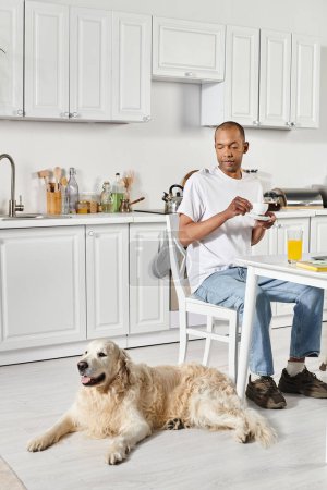 Téléchargez les photos : Un Afro-Américain handicapé assis à une table de cuisine avec son fidèle chien Labrador à ses côtés. - en image libre de droit