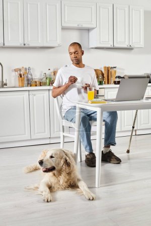 A disabled African American man collaborates with his loyal Labrador dog while working on a laptop at a table.