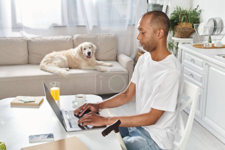 A disabled African American man sit at a table using a laptop computer, accompanied by a Labrador dog.