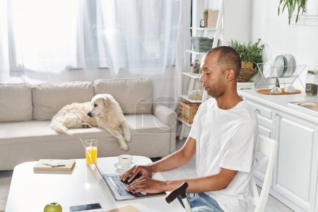 Ein behinderter Afroamerikaner sitzt mit einem Laptop an einem Tisch, begleitet von seinem Labrador-Hund.