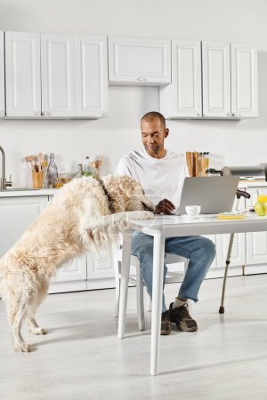 A disabled African American man sits at a table with a laptop open in front of him, accompanied by his loyal Labrador dog.