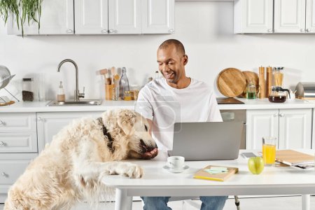 A disabled African American man sits at a table with a laptop, accompanied by his loyal Labrador retriever.