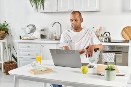 Un hombre afroamericano diverso con miastenia gravis se sienta en una mesa de cocina, absorto en su computadora portátil.