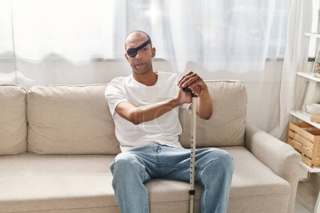An elderly African American man with myasthenia gravis sits on a couch with a cane, lost in thought.