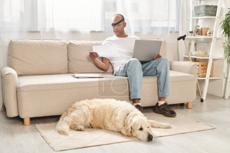 Téléchargez les photos : Un homme, luttant contre la myasthénie grave, s'assoit sur un canapé avec un ordinateur portable, accompagné de son fidèle chien Labrador. - en image libre de droit