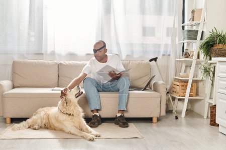 A disabled African American man with myasthenia gravis syndrome sits on a couch next to his Labrador dog.