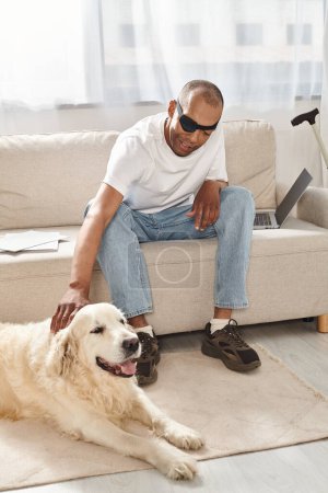 Foto de Un hombre afroamericano discapacitado con síndrome de miastenia gravis se sienta junto a un perro leal Labrador en un sofá. - Imagen libre de derechos