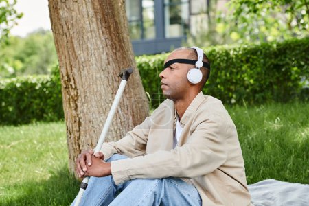 A man with headphones is seated on a blanket next to a tree, enjoying music and the peaceful surroundings