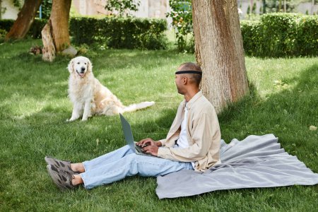 Ein behinderter Afroamerikaner mit Myasthenia-Gravis-Syndrom sitzt mit einem Laptop im Gras, begleitet von einem Labrador-Hund.