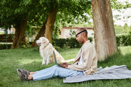 A diverse man with Myasthenia Gravis sits on the grass, using a laptop while accompanied by his loyal Labrador dog.