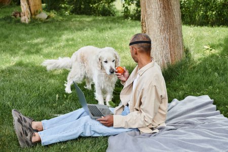 An African American man with myasthenia gravis sits in grass with his laptop while a Labrador dog stays by his side.
