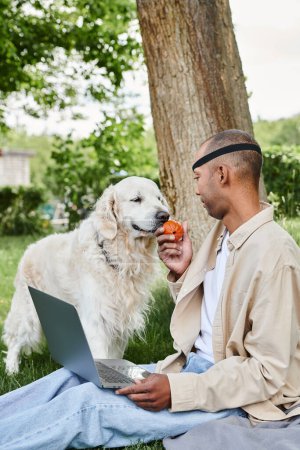 Téléchargez les photos : Un homme atteint du syndrome de la myasthénie grave, assis dans l'herbe, travaillant sur un ordinateur portable à côté de son fidèle chien Labrador. - en image libre de droit