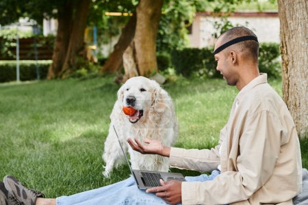An African American man with myasthenia gravis sits on grass with a laptop, balancing a ball in his mouth as his Labrador looks on.