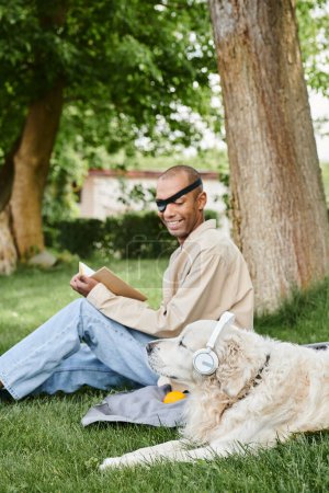 Téléchargez les photos : Un homme atteint du syndrome de la myasthénie grave est assis avec son chien du Labrador dans l'herbe, tous deux portant un casque d'écoute. - en image libre de droit