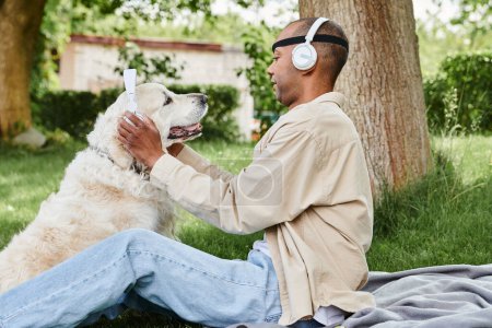 A disabled African American man with myasthenia gravis syndrome sits in the grass with a Labrador dog wearing headphones, enjoying a peaceful moment together.