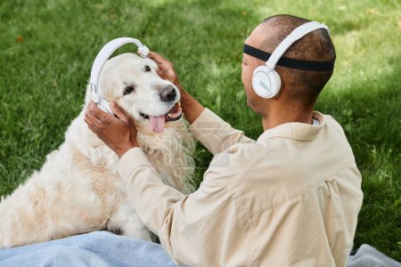 A diverse man with myasthenia gravis syndrome sits on grass, joyfully petting his loyal Labrador dog while both wear headphones.