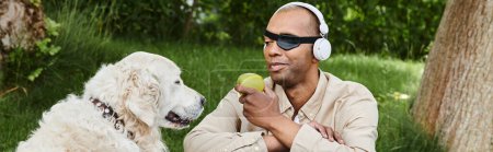 Ein behinderter Afroamerikaner mit Myasthenia-Gravis-Syndrom hört Kopfhörer, während er neben seinem treuen Labrador-Hund einen Apfel isst.