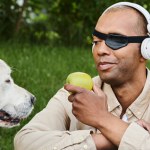 A disabled African American man with myasthenia gravis syndrome listens to headphones while eating an apple beside his loyal Labrador dog.