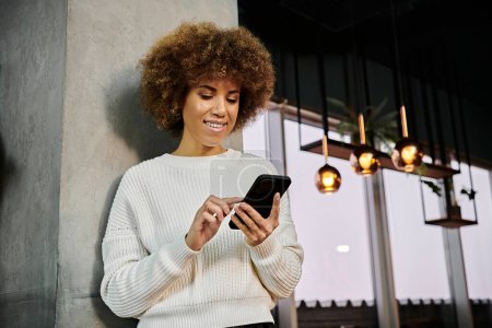 An African American woman in a white sweater captivated by her cell phone in a modern cafe setting.