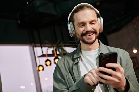 Foto de Un hombre, auriculares encendidos, sosteniendo un teléfono celular, perdido en la música y la conversación en un moderno entorno de cafetería. - Imagen libre de derechos