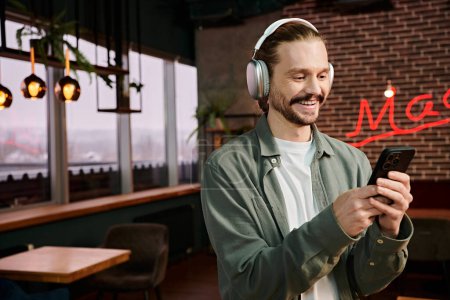 A man, wearing headphones, gazes at his phone in a modern cafe, immersed in his own world of music.