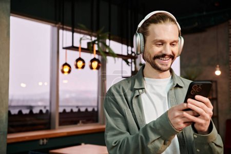 A man, wearing headphones, gazes at his phone in a modern cafe while connected to music.