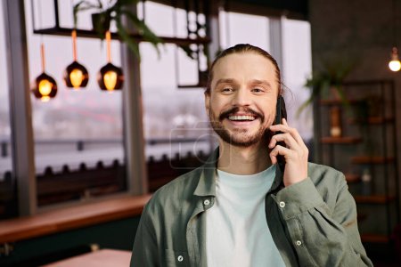 man with a stylish beard talking animatedly on a cell phone in a modern cafe setting.