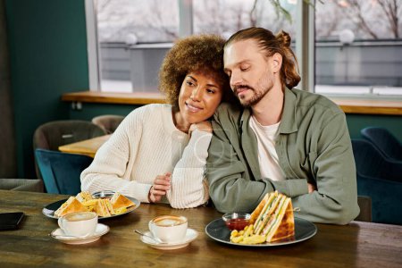 Une femme afro-américaine et un homme sont assis à une table, partageant un repas dans un café moderne.