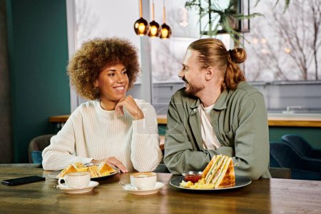 Una mujer afroamericana y un hombre se sientan en una mesa cargada de platos de comida en un café moderno, disfrutando de una comida juntos.
